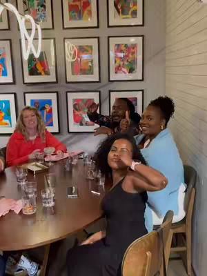 A group of people are seated around a round dining table, engaged in conversation. The setting is a well-lit room with framed artwork on the wall behind them. The composition focuses on the interactions and the atmosphere of the dining experience.