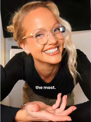 A close-up shot of a young woman with fair skin, blonde hair styled in a loose updo with face-framing strands, and large-framed glasses. She is smiling broadly, revealing perfectly aligned white teeth, with a warm, engaging expression. Her attire includes a black top, and her hands are visible, resting near the lower portion of the frame. The background is slightly blurred, keeping the focus on the subject.
