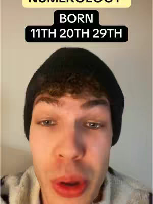 A selfie-style video cover. The creator is centered, with their face filling most of the frame, set against a plain, light-colored background. The creator wears a black beanie and a light colored shirt. Above the creator's head are two text boxes: 'NUMEROLOGY' in a yellow rectangle, and below it a black rectangle with white text that reads 'BORN 11TH 20TH 29TH'.