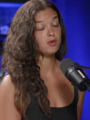 A woman with long, wavy brown hair is positioned slightly off-center. She is wearing a black top and has a microphone in front of her, suggesting a recording or speaking scenario. The background is a solid dark blue. Her eyes are closed, and her mouth is slightly open, as if she is singing or speaking. The overall composition is simple, focused on the person's expression.