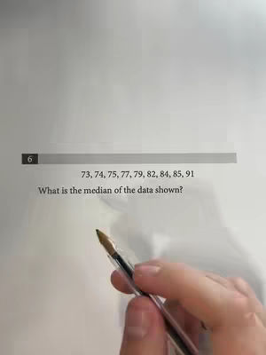 A cropped image shows a textbook page with a math problem about finding the median of a data set. A pen is visible in the bottom right, held by a person whose hand is partially visible.