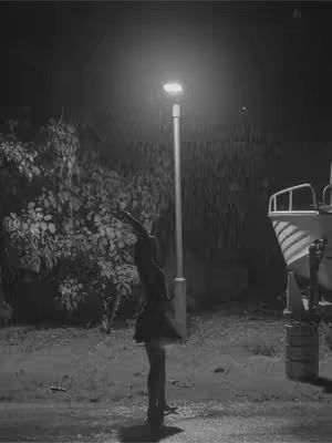A black and white photo shows a woman wearing a dress standing in a dark outdoor setting next to a street lamp. The woman has one arm up in the air. Behind her are some leafy features, next to a boat-like structure.