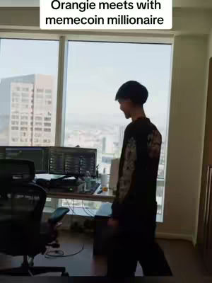 A man stands in a well-lit office setting near a large window showing a cityscape. He is turned away from the camera and is wearing dark clothing. A desk with computer monitors is visible, suggesting a trading or office environment. The text overlay indicates a meeting with a meme coin millionaire.