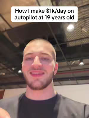 A selfie-style video cover image features a young man in a black shirt, centered in the frame. The background appears to be an indoor setting with visible lighting and architectural elements. The man is looking at the camera with a slightly inquisitive expression. Above him, a text overlay states: "How I make $1k/day on autopilot at 19 years old".
