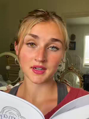 A woman with blonde hair and two different colored eyes is holding a book and looking at the camera. Her lips are parted as if she is talking. She is wearing a red top over a gray one.