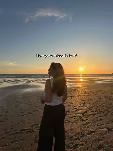 A woman stands on a beach at sunset, facing away from the camera. The sky is a gradient of blues and oranges, reflecting onto the wet sand. Text overlayed reads 'Have you ever been in love?'