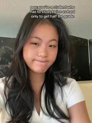 A selfie-style shot of a young woman with long, dark hair and a white top. She's looking at the camera with a slightly pensive expression, lips pressed together. The background is a simple, slightly blurred interior.