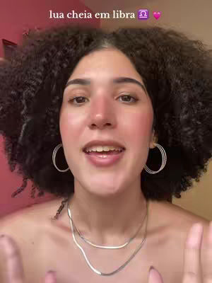 A woman with curly dark hair and fair skin is looking at the camera. She's wearing hoop earrings, a layered necklace, and has a soft, natural makeup look. The text overlay indicates astrological themes.