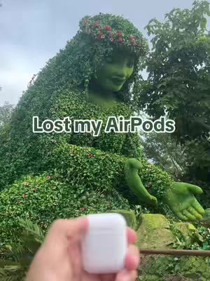 The cover image shows a person holding a white AirPod case in front of a statue of a woman covered in meticulously trimmed green plants. The woman statue has floral decor on the head, the background contains trees and the sky is visible. The text overlay on the image says 'Lost my AirPods'.
