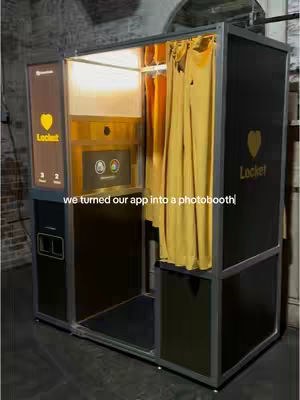 An image of a photo booth with a custom design, branded with the 'Locket' logo. The booth is primarily black and metallic silver with accents of yellow, and the background appears to be a brick wall. Yellow curtains are seen inside the booth.