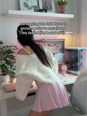 A woman is shown from the back, standing near a desk in front of a computer. The woman is wearing a white off-the-shoulder sweater and a pink skirt. She has long black hair. On the desk, there's a pink computer setup with a monitor, a pink keyboard and mouse, and a pink computer tower. A shelf with picture frames, plants, and decorations is behind her. The overall scene has soft pink and white hues, giving a cozy aesthetic.
