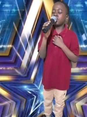 A young boy wearing a red polo shirt and tan pants stands center frame, holding a microphone. He's singing on a stage with a background composed of a blue-and-violet star-shaped design. The image is well-lit with the focus on the boy, capturing a moment of performance.