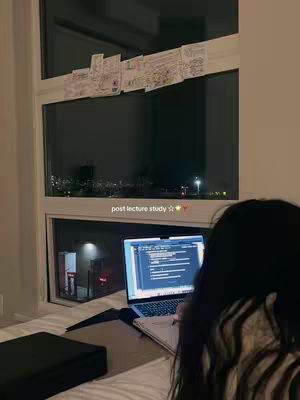 The cover image shows a person studying at night. The scene is set in a room with a large window overlooking a city at night, suggested by the lights in the dark background. The person, seen from the back, is sitting on a bed near the window, focused on a laptop. Colorful notes are taped to the window frame. The overall composition is dimly lit, with a warm color palette.