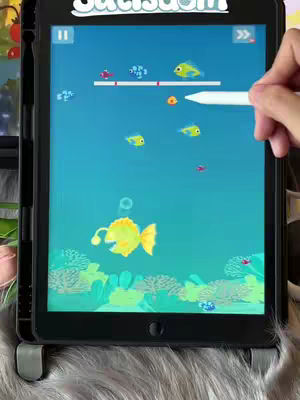 A tablet displaying a mobile game. The screen shows an underwater scene with animated fish, bubbles, and coral. A hand with a stylus hovers over the screen.
