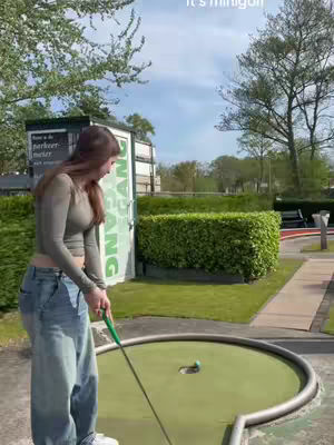 A woman in casual wear prepares to putt a golf ball on a miniature golf course. Lush green foliage and a bright sky serve as a backdrop.