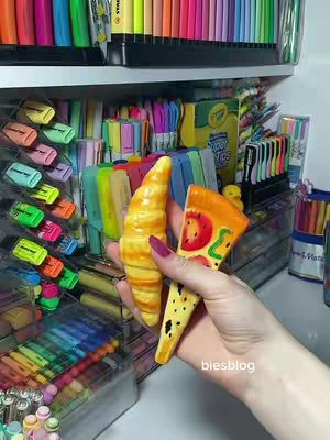 A person's hand holds a croissant and a pizza slice in front of a shelf filled with colorful stationery items such as pens and highlighters. The background is filled with various office supplies, creating a vibrant and organized setting.