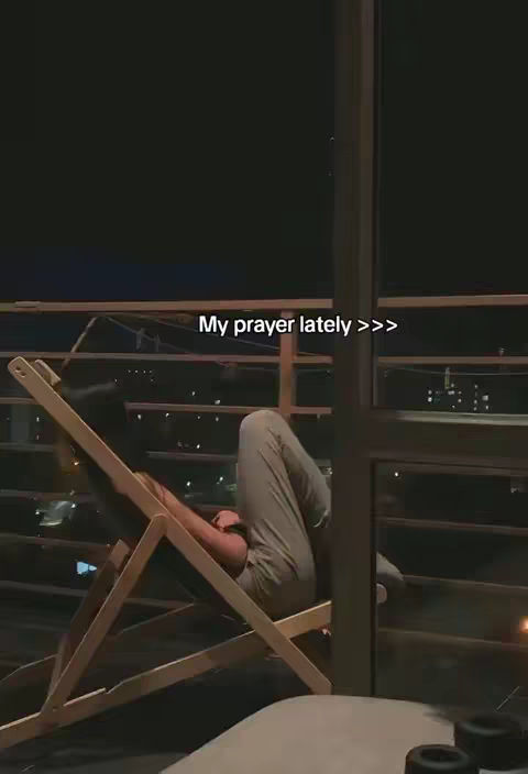 A medium shot shows a person upside down, leaning back in a wooden chair on a balcony. The person is wearing light-colored clothing. The background is a dark night scene with city lights visible in the distance. The text overlay states, "My prayer lately >>>".