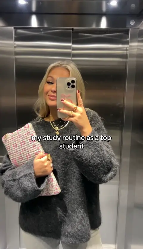 A woman is holding her phone up to take a selfie in an elevator. The elevator walls are stainless steel, and she is wearing a dark gray fluffy sweater. In the image, there is text in the lower portion; the woman is smiling and holding a pink and white patterned pouch. She is also wearing multiple rings, necklaces, and the nails are red.