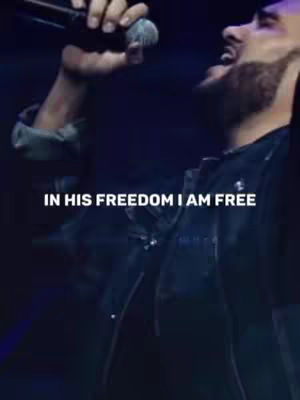 A man is singing with a microphone in his hand. Text overlay says 'IN HIS FREEDOM I AM FREE.' The background is dark, creating a spotlight effect on the man.