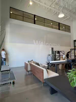 An interior shot of a large, modern space. A white wall dominates the scene, with the text of the Bible subtly printed across. A long, dark-framed window runs along the top of the wall. Below, a low-profile sofa with a wooden base and cream-colored cushions is placed. Various pieces of equipment, including a large printing machine, are scattered throughout the open area, suggesting a church or religious facility engaged in a project. Other individuals are present, working or observing, near a table and additional equipment.
