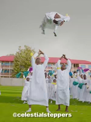 The image shows a group of people dressed in white robes, dancing and celebrating outdoors. One person is in the air mid-flip. The setting appears to be a sunny, grassy area with a building in the background.