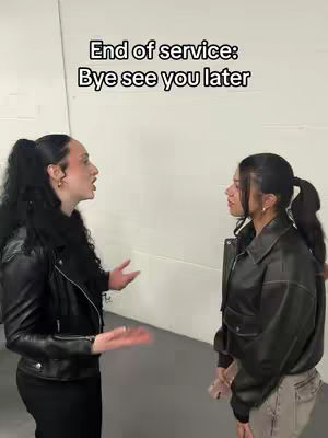 Two young women are positioned in a room with plain white walls. The woman on the left is talking with her hands open, while the woman on the right is listening.