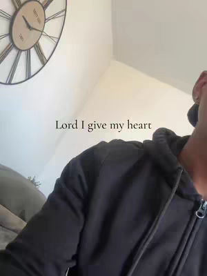 The image features a person wearing a black hoodie, with their face partially visible, set against an interior background. A clock is mounted on the wall and the person is holding the camera from the side. The text overlay reads "Lord I give my heart".
