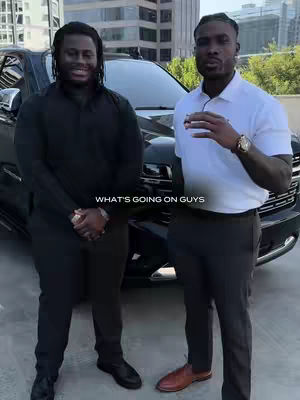 Two men standing in front of a black SUV. The man on the left is wearing a black outfit and smiling slightly. The man on the right is wearing a white polo shirt and appears to be talking.