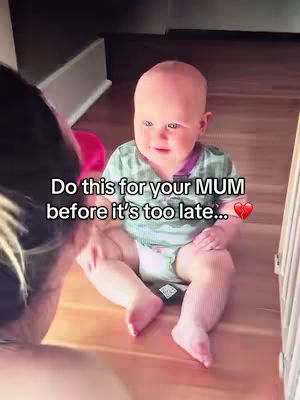 A baby is seated on a wood floor, viewed from a high angle. Text in white font overlays the image, with the text 'Do this for your MUM before it's too late...' and a broken heart emoji. The baby is gazing towards the viewer. A person is seen in the over-the-shoulder view on the left.