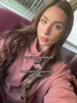 A selfie-style shot featuring a woman with long, wavy brown hair, wearing a pink top. Text overlays divide the screen, contrasting astrological signs: 'cancer/scorpio/pisces men vs. capricorn/taurus/virgo women.' The background is of an indoor setting, with a blurry view of what appears to be a window overlooking a city.
