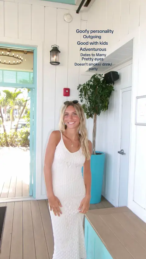 A woman in a white dress stands facing the camera indoors. The background shows a white wall and a doorway with a view of trees. Text overlays describe the woman's characteristics.