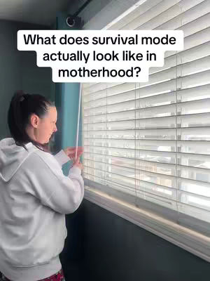 A woman in a white hoodie stands next to a window with closed blinds. The image has a text overlay in white. The setting appears to be indoors, with the wall beside the window painted in a dark teal.