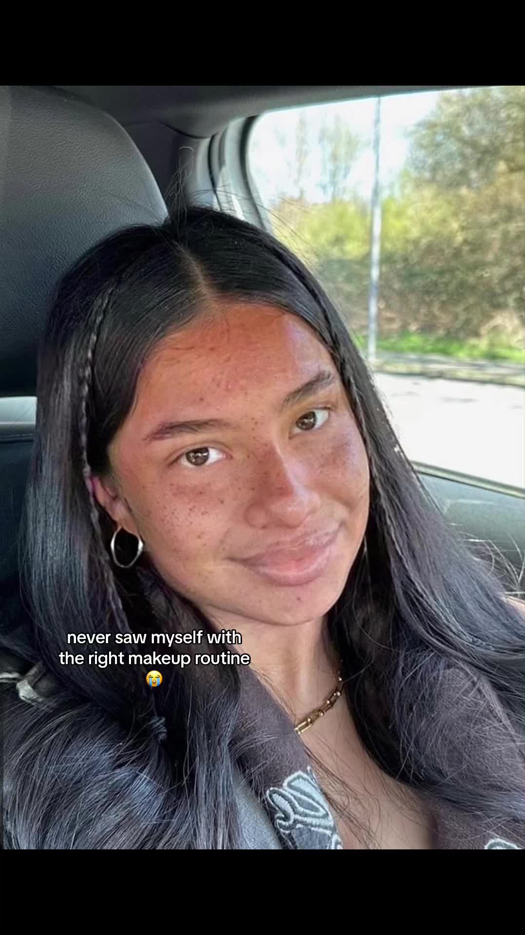 The image shows a close-up of a person in a car, likely taking a selfie. The person has dark hair partially pulled back, fair skin with noticeable freckles, and is wearing hoop earrings. The background shows a blurred view of greenery, suggesting an outdoor setting. Text overlayed on the image reads, 'never saw myself with the right makeup routine'.