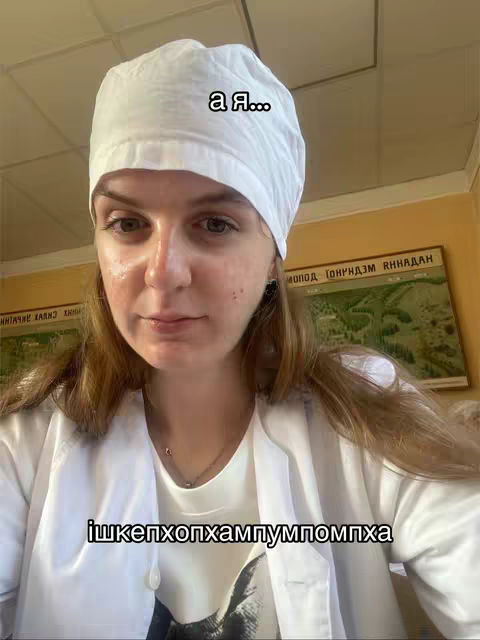 A selfie-style shot featuring a young woman dressed in medical scrubs and a surgical cap. She is looking directly at the camera with a mild expression. The background shows details of what seems to be an office or a study room with some documents or posters on the wall.