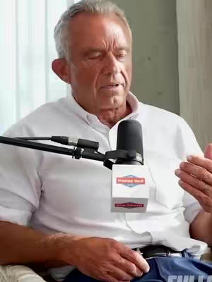 A man with short gray hair and a white button-down shirt is seated, with a microphone in front of him. The background includes a window with light-colored curtains. The man is gesturing with his right hand while speaking, the microphone is positioned with the words "Happy Dod" in white text on a red rectangle.