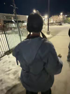 A person is walking away from the camera along a snowy sidewalk next to a metal fence, with buildings and streetlights in the background at night.