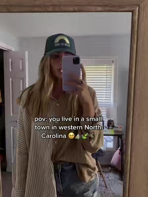 A woman stands in front of a mirror, holding her phone to take a selfie. She wears a baseball cap, a button-up shirt over a t-shirt, and jeans. Text overlaid on the image reads 'pov: you live in a small town in western North Carolina'. The background reveals a tidy bedroom with a window and a desk.