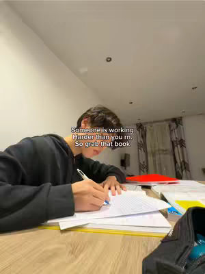 A person is sitting at a desk, writing on a piece of paper. The background is a bit out of focus, but there is a book and a bag on the desk, and the person is wearing a black hoodie. The text overlay says 'Someone is working Harder than you rn. So grab that book'.