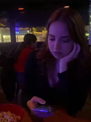 A woman with auburn hair sits, resting her chin on her hand, looking down at a phone. She is in a dimly lit setting, likely a bar, with another person partially visible in the background, and a bowl of snacks visible on the table.