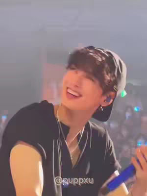 The image shows a young man wearing a black cap and shirt, smiling broadly with his mouth open. The background seems to be a stage setting, with a blurred audience and lighting, suggesting a performance or concert scene. He appears to be holding a microphone.