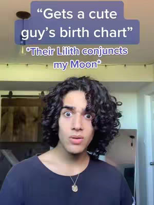 A young man with dark curly hair and wide eyes is the central focus. He is wearing a dark blue t-shirt and a gold necklace. Above him is a blue rectangle with white text, providing context for the expression he's making. The background, though slightly out of focus, suggests an interior setting with neutral-colored walls and what seems to be a partially visible mirror.