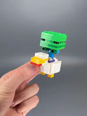A hand holds a Lego creation that appears to be a Minecraft character riding a chicken. The head of the rider is green, made of Lego bricks. The chicken/mount is white, with yellow and red details. The background is a plain, grey surface.