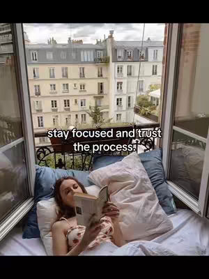 A woman lies in bed, propped up by pillows, reading a book. She's positioned near an open window overlooking a European cityscape.
