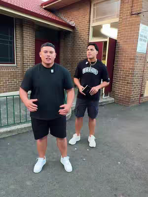 Two young men stand outside a building, likely a church, in the cover image for a music video. The man on the left is the primary focus, wearing a black t-shirt and shorts, and appears to be gesturing with his hands. The man on the right stands slightly behind him, also in black, holding a small black object. The setting has a warm, overcast light, giving the image a natural feel. The image shows a brick building in the background with a red roof and a large window. The image suggests music and religious themes.