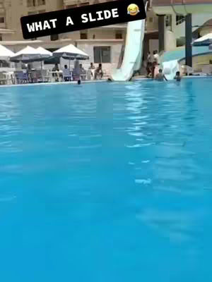 A wide shot of a swimming pool area, featuring a large pool of bright blue water. To the right, a water slide is visible. The image is bright and sunny, suggesting an outdoor setting. In the center, a black text overlay reads "WHAT A SLIDE" with a smiling emoji.