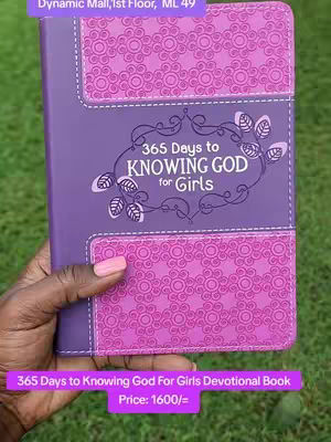A hand holds an open book with a purple cover. The background is green grass. The book's cover is purple and features a floral design in a lighter shade of purple on its top and bottom portions. In the center, there's a decorative frame in a darker purple color, containing the text '365 Days to Knowing God for Girls' in a white, elegant font. The price and bookstore location are displayed in a purple bar. The book is the main focus, displayed against a natural backdrop.
