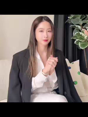 A woman with long brown hair is centered in the frame. She wears a white button-down shirt and a black blazer. Her hands are clasped in front of her. The background includes a neutral wall, a dark curtain, and part of a leafy plant and a decorative pillow on the couch.