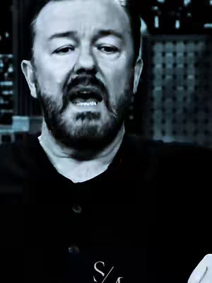 A black and white image of Ricky Gervais speaking, with his mouth open. He has a full beard and is wearing a dark shirt. The background appears to be part of a set or stage.