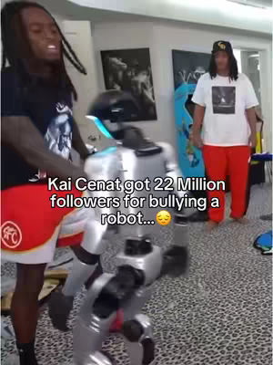 The image captures a video of a man interacting with a metallic robot. The man, on the left, is Black, wearing a black t-shirt and red shorts, looking animated. Next to him, the robot is silver with blue lights. In the background, another individual in red pants and a white t-shirt stands, wearing a cap. The setting is a brightly lit room with a patterned floor and artwork on the walls.