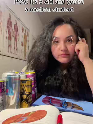 A woman with long, dark, wavy hair sits at a table, looking tired. She wears dark-rimmed glasses and her face is illuminated by what looks like a ring light. The table is cluttered with energy drink cans, textbooks, and a tablet showing a medical diagram. Behind her, an anatomy chart hangs on the wall.